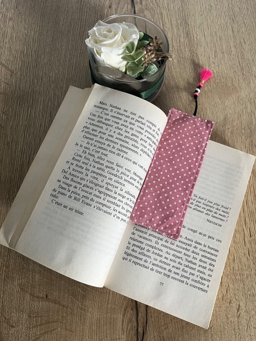 Marque-page en tissu rose à pois posé sur un livre ouvert, avec cordon et pompon rose dépassant en haut.