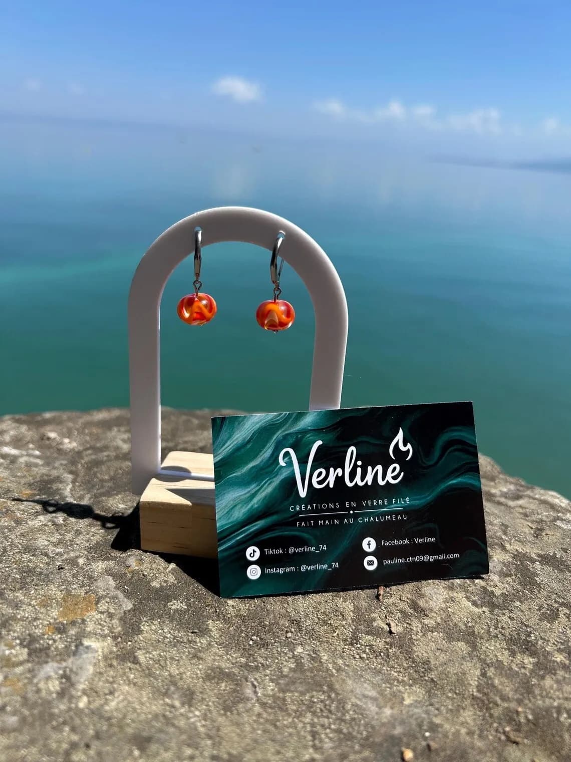 Boucles d'oreilles avec perles en verre filé orange et ambre sur un support blanc face à la mer.